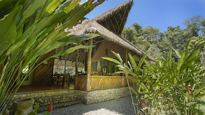 Uma construção com telhado de palha e base de pedra, rodeada por vegetação tropical.