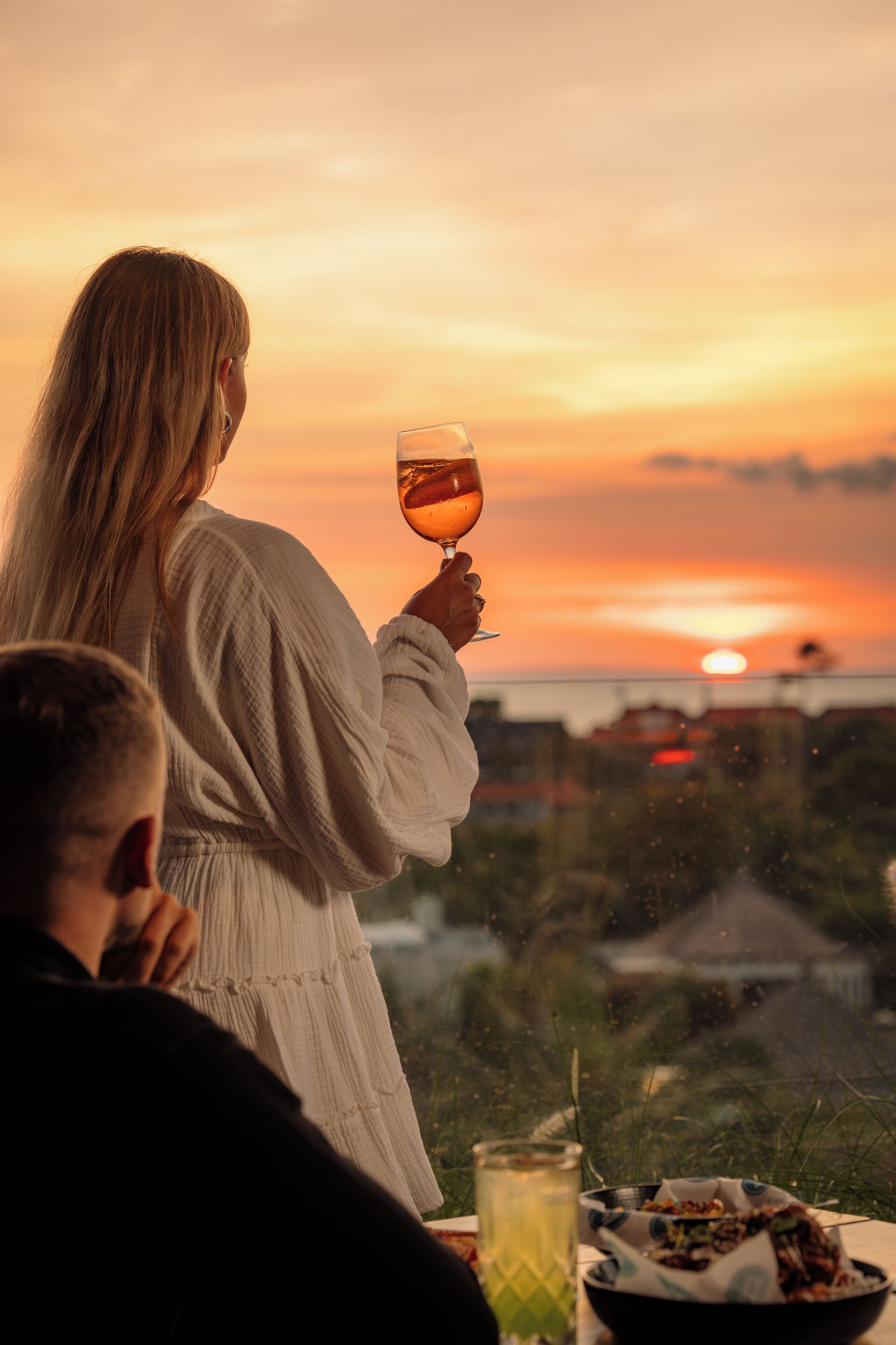 Une personne tenant un verre de vin, un bol de nourriture et une boisson sur une table, avec une vue sur le coucher de soleil.