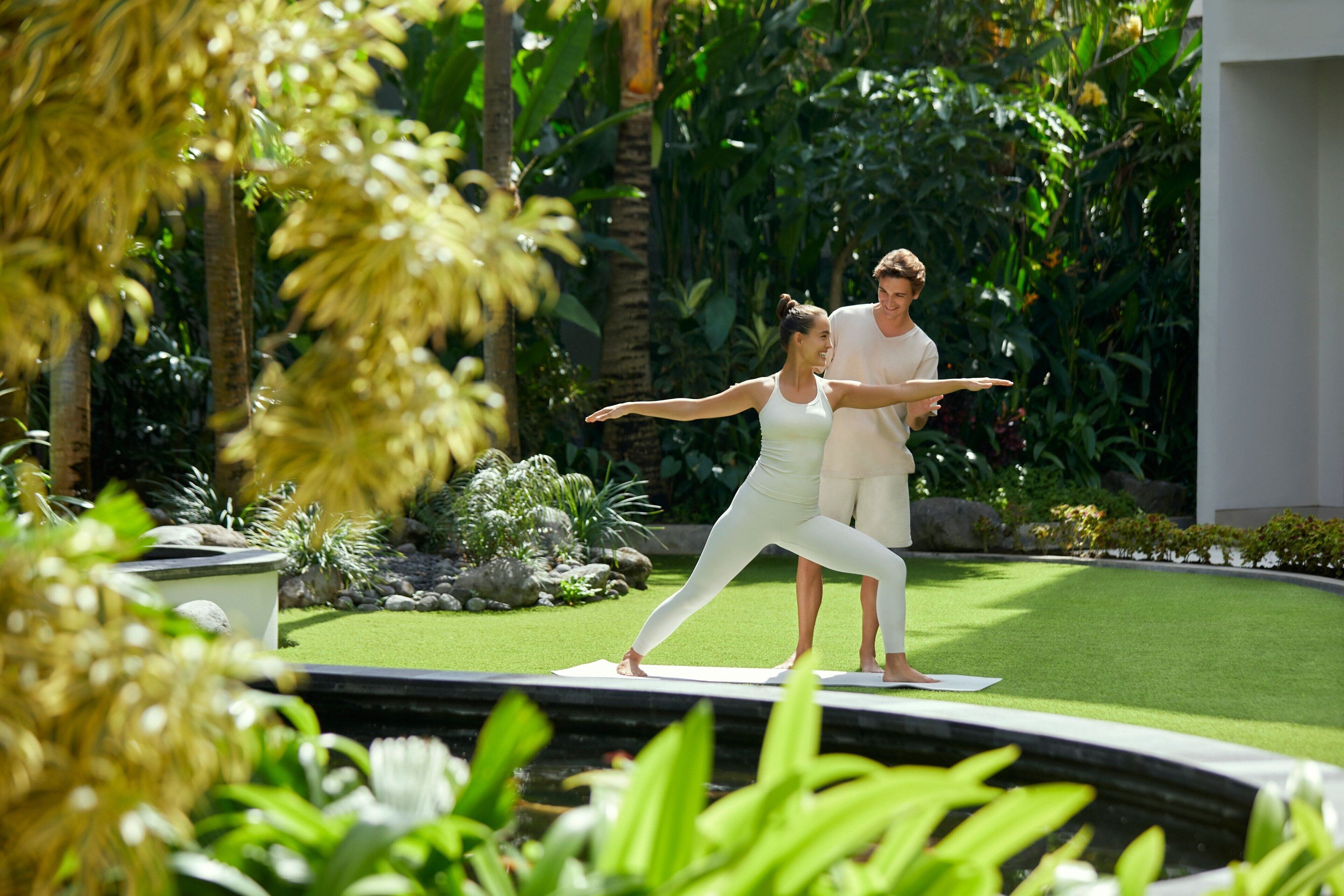 A couple practicing yoga on a white mat in a lush garden with tropical plants and a small pond.