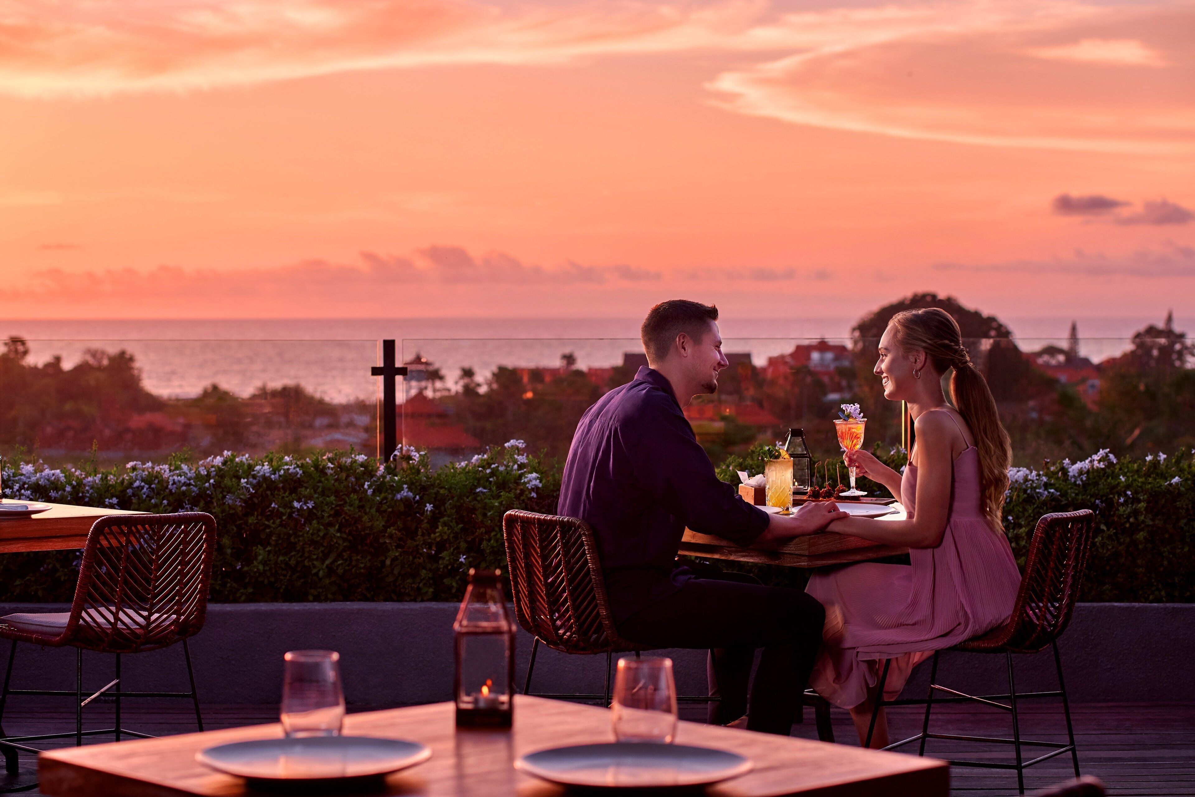 A couple dining at an outdoor table with a view of the ocean and cityscape during sunset.