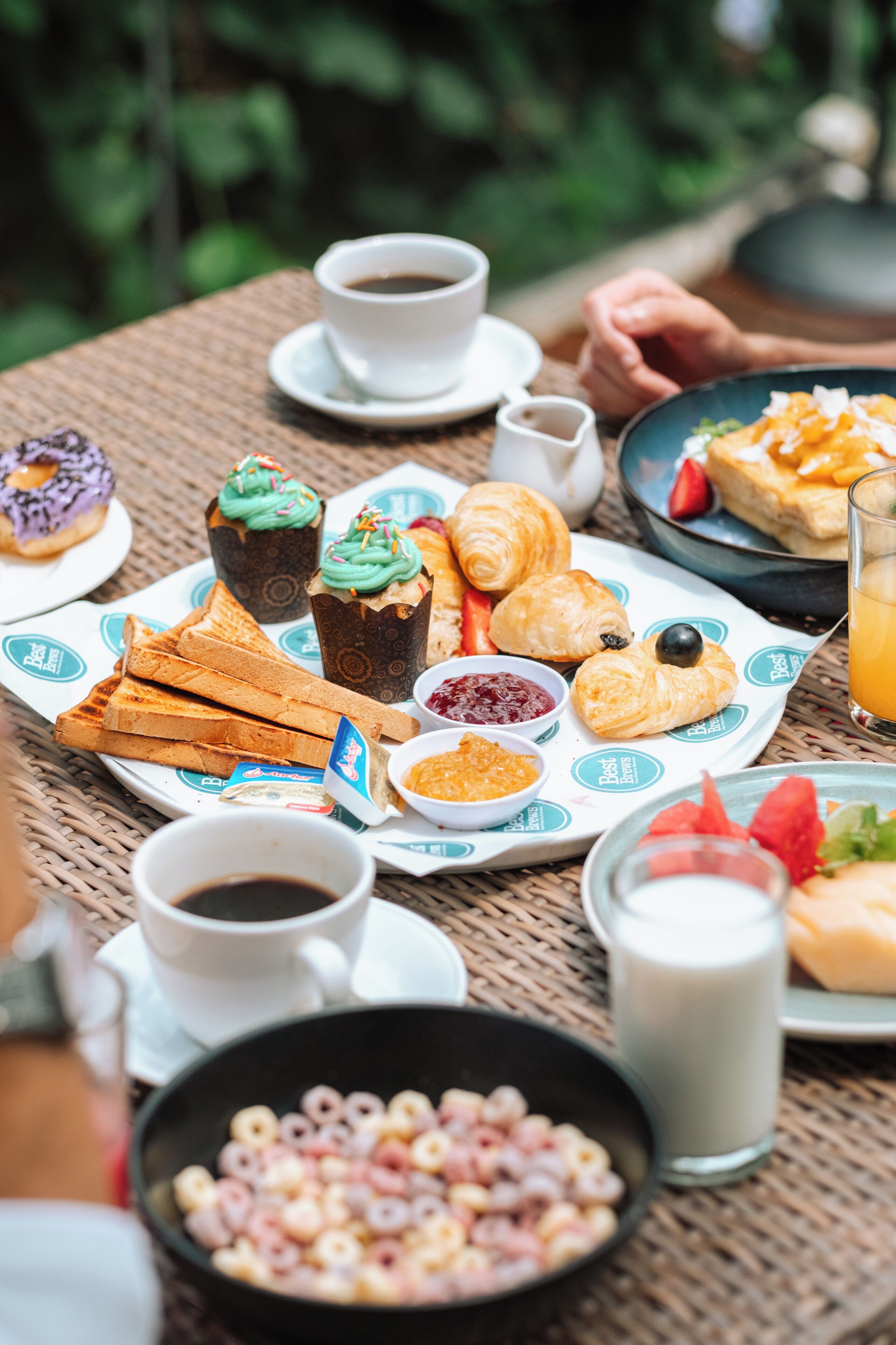 Un assortiment de produits pour le petit-déjeuner, disposé sur une table en osier, comprenant des croissants, des viennoiseries, des fruits et diverses boissons.