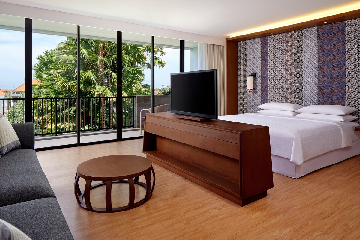 Ein modernes Hotelzimmer mit einem großen Bett, einer Couch, einem Fernseher auf einer Holzkonsole und einem Balkon mit Blick auf Palmen.