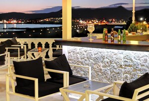 A terrace with outdoor seating, a bar area with bottles, and a view of a cityscape at dusk. - The Royal Hotel (Corfu)
