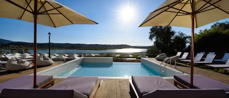 A poolside area with lounge chairs and umbrellas, a wooden deck, and a view of a lake and mountains.