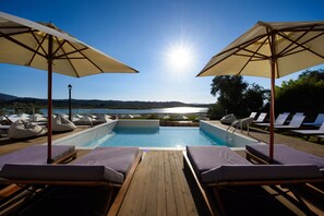 A poolside area with lounge chairs and umbrellas, a wooden deck, and a view of a lake and mountains. - The Royal Hotel (Corfu)