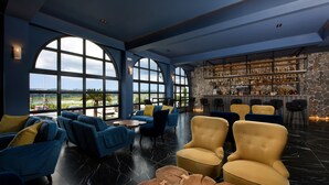 A spacious lounge area with large windows, comfortable seating, and a bar with a stone wall backdrop. - The Royal Hotel (Corfu)