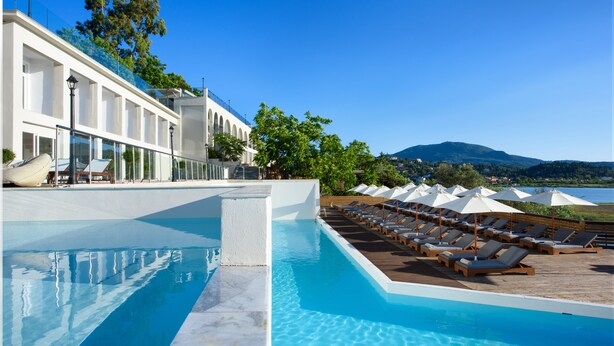 A pool area with lounge chairs and umbrellas, a white building with arched windows, and a view of a mountain. - The Royal Hotel (Corfu)
