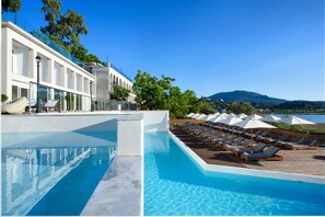 A pool area with lounge chairs and umbrellas, a white building with arched windows, and a view of a mountain. - The Royal Hotel (Corfu)