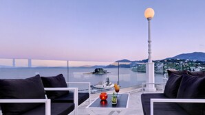 A terrace with black cushioned seating, a glass table with a drink and candle, and a view of a bridge and water. - The Royal Hotel (Corfu)