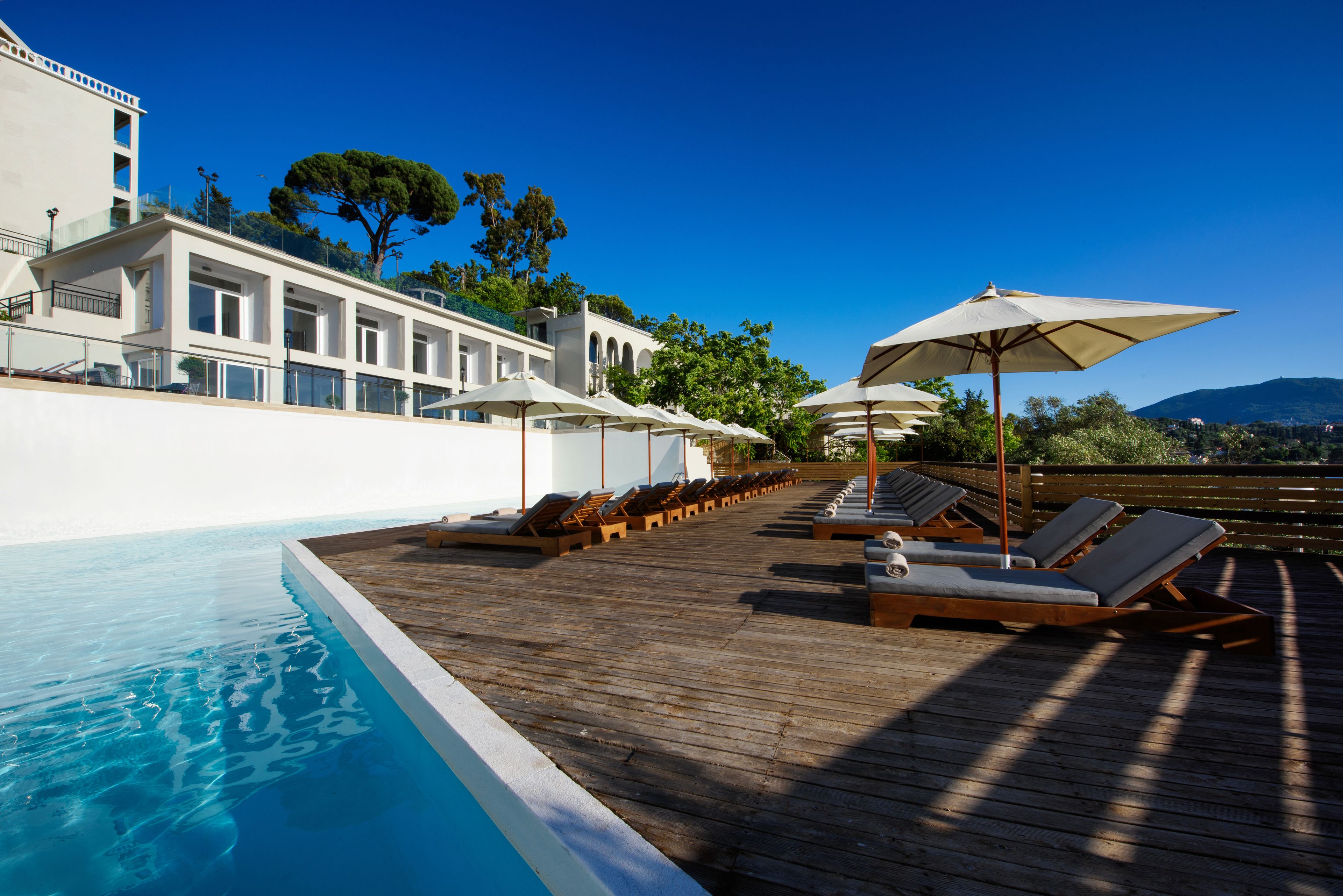 Terraza de madera con sillones y sombrillas, una piscina y un edificio con balcones.
