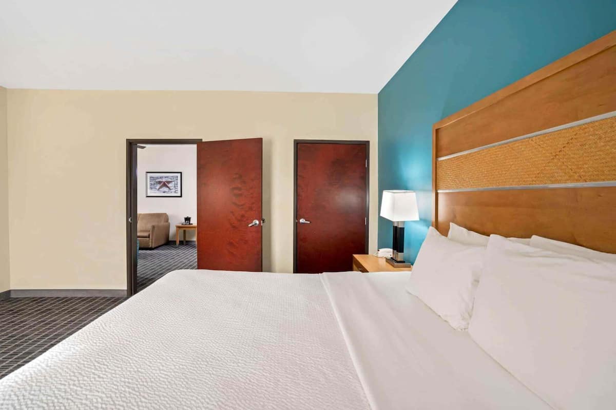 A hotel room with a large bed, two doors, a nightstand with a lamp, and a small table with a chair.