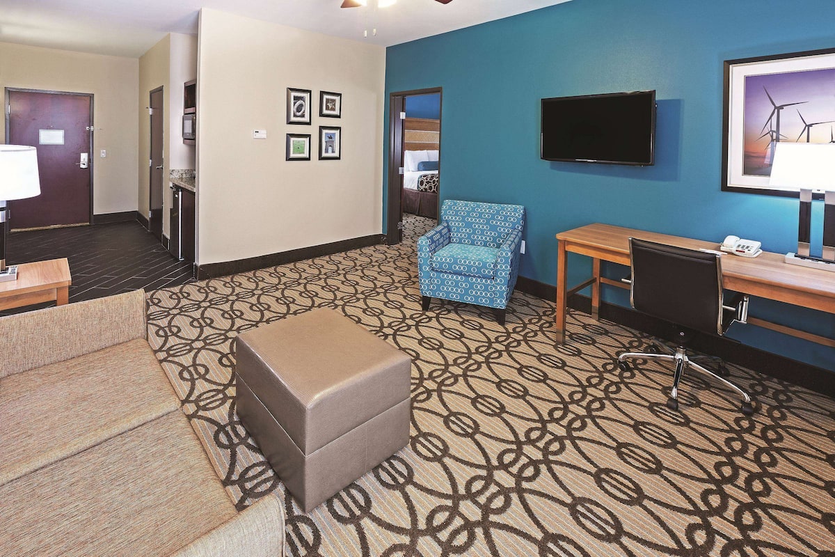 A hotel room with a patterned carpet, a sofa, a small ottoman, a desk with a chair, a television, and framed pictures on the wall.
