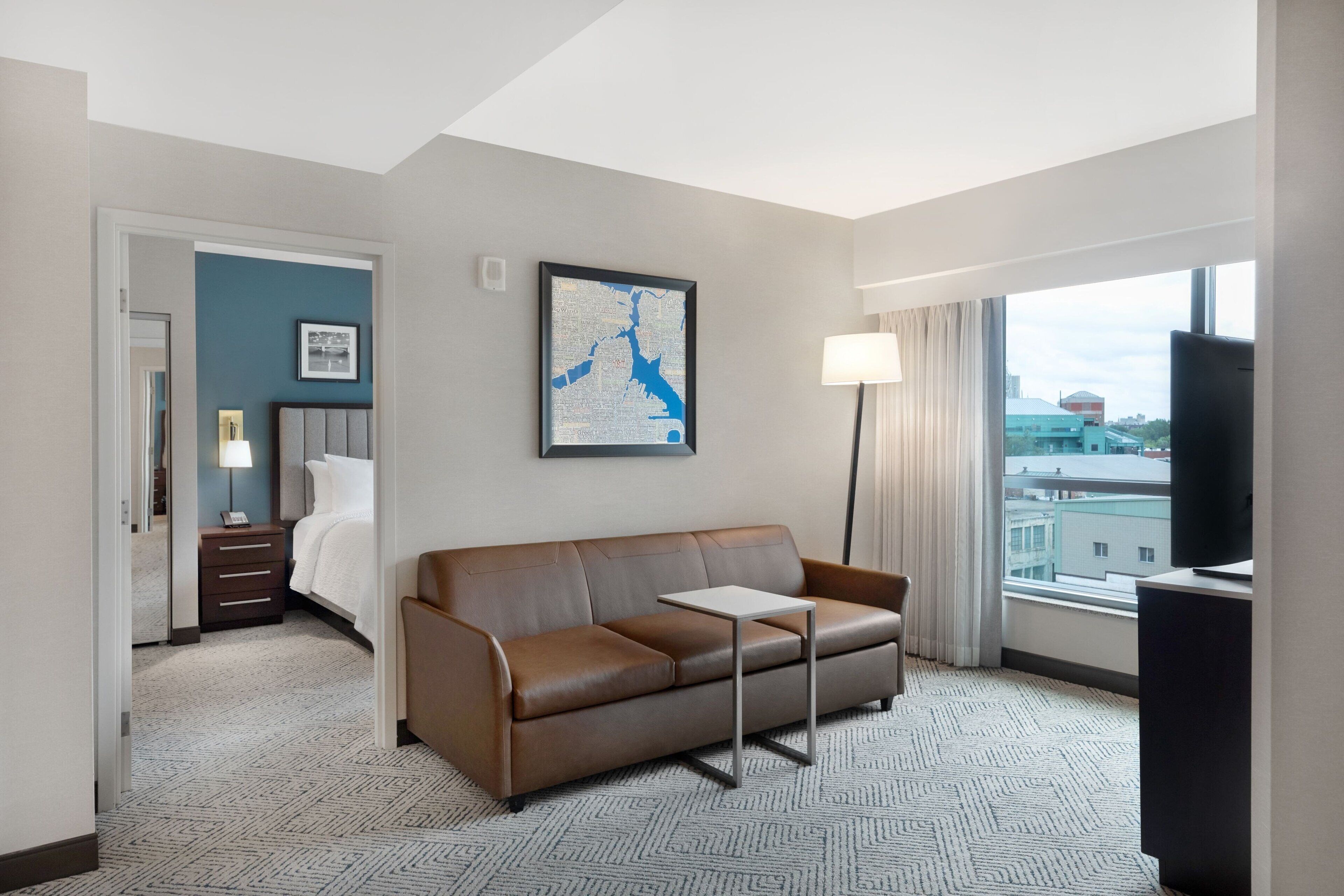 Habitación de hotel con sofá de cuero marrón, una mesita, cama con sábanas blancas, un mapa en la pared y vistas a edificios a través de la ventana.