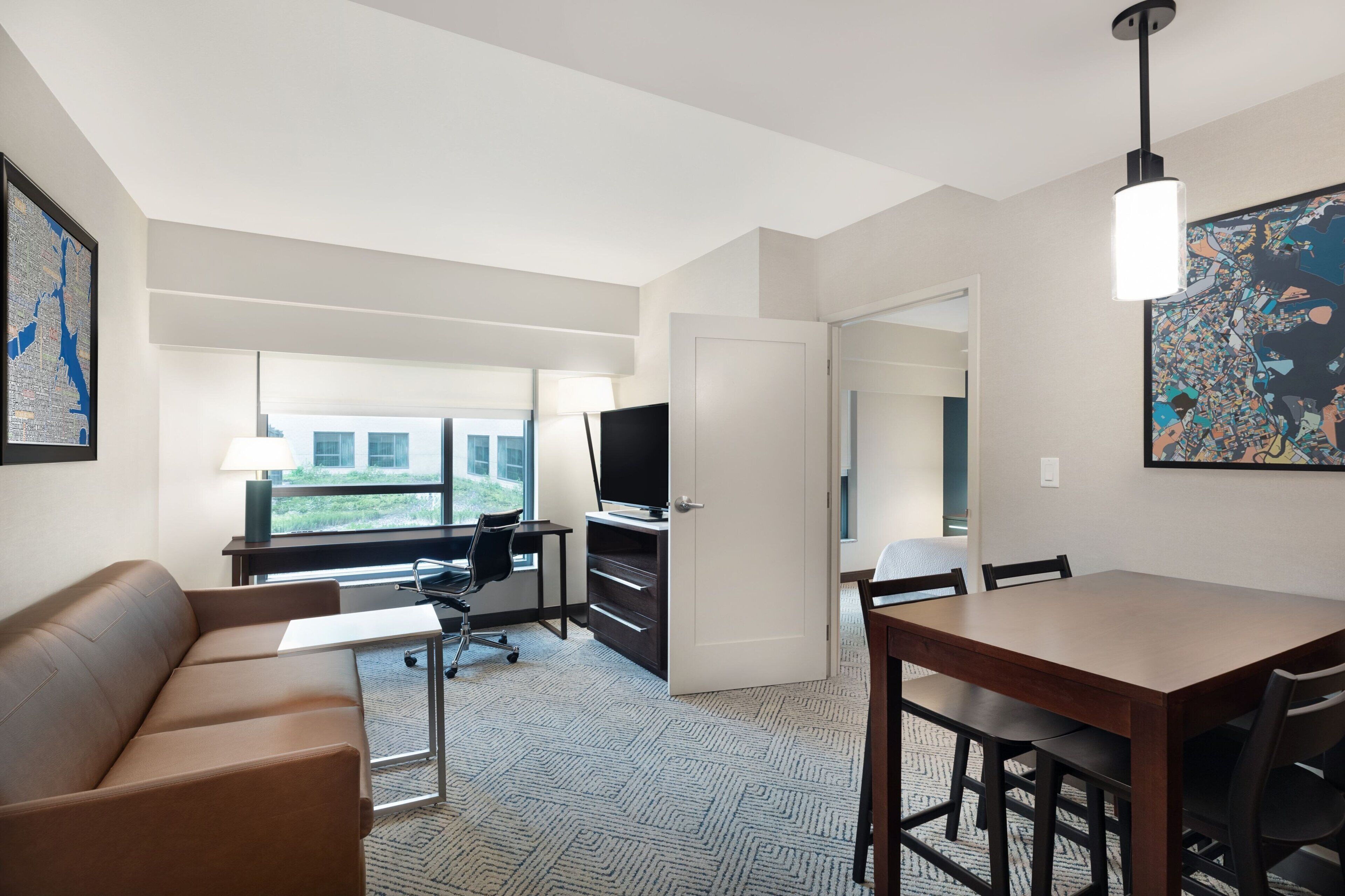 A modern hotel room with a sofa, a desk with a computer, a TV, a dining table with chairs, and artwork on the walls.