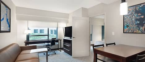 A modern hotel room with a sofa, a desk with a computer, a TV, a dining table with chairs, and artwork on the walls.