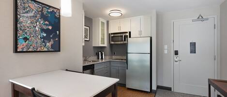 A compact kitchen with white cabinets, a stainless steel refrigerator, and a microwave. There's a dining table with chairs and a hanging light fixture.