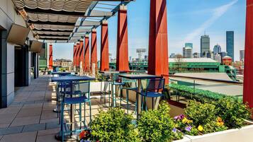 An outdoor seating area with tables and chairs, a view of the city skyline, and a green-roofed building in the background.