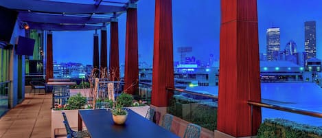 Una terraza al aire libre con una mesa y sillas, vista al horizonte de la ciudad y columnas estructurales rojas.