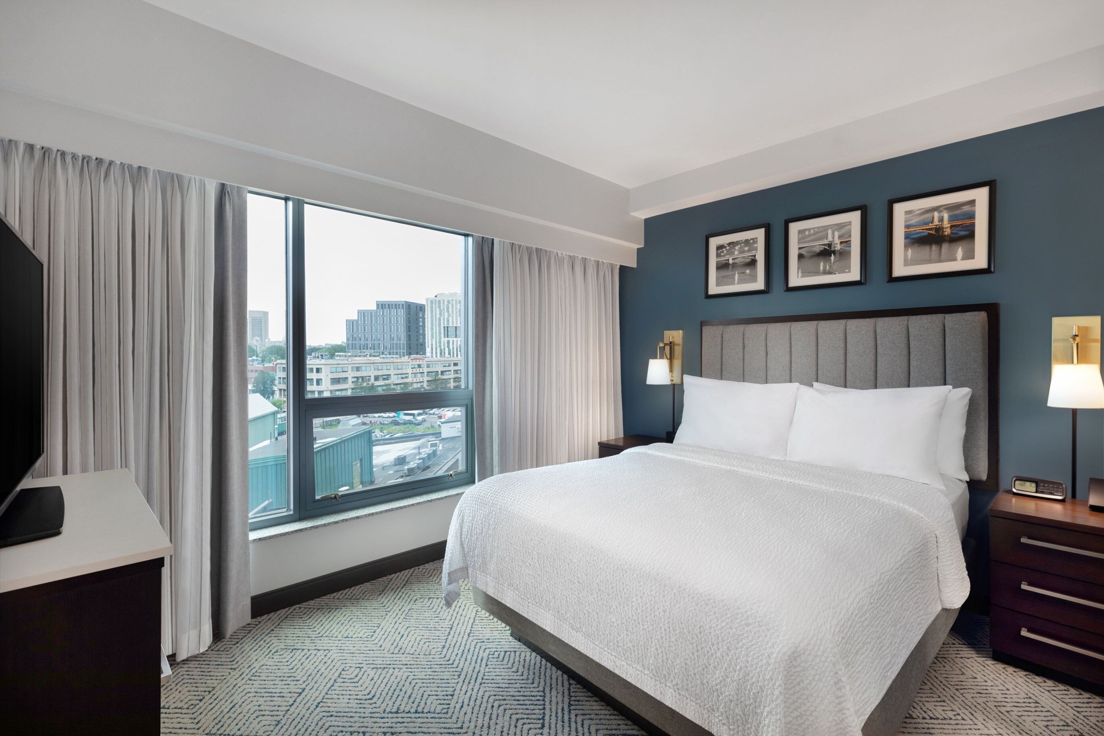 A hotel room with a large bed, a TV on a desk, a window with curtains, and framed pictures on the wall.