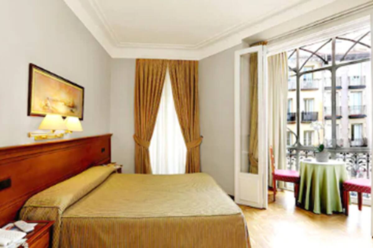 A hotel room with a bed, a desk, a chair, a table with a green tablecloth, and a view of the outside through a window with curtains.