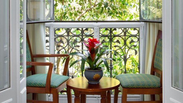 Un balcón con una mesa y sillas, una planta en maceta con flores, y vistas a un edificio y árboles.