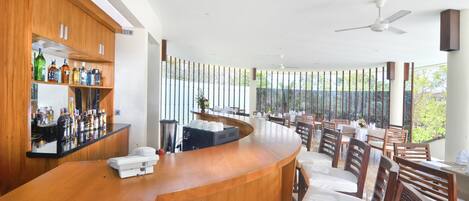Un espace bar moderne avec des meubles de rangement en bois et un comptoir incurvé. On y trouve plusieurs coins salon avec des tables et des chaises, ainsi que de grandes fenêtres offrant une vue sur la verdure environnante.