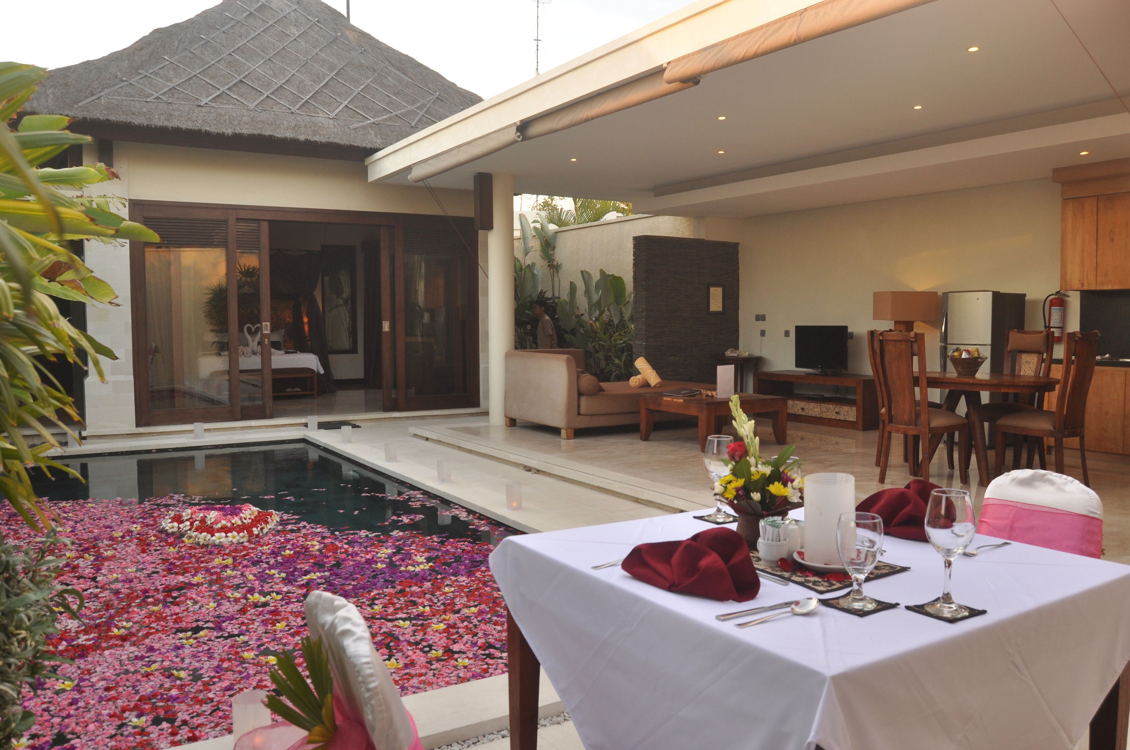 A dining area with a poolside setting, featuring a table set for two with a white tablecloth, red napkins, and a vase of flowers.