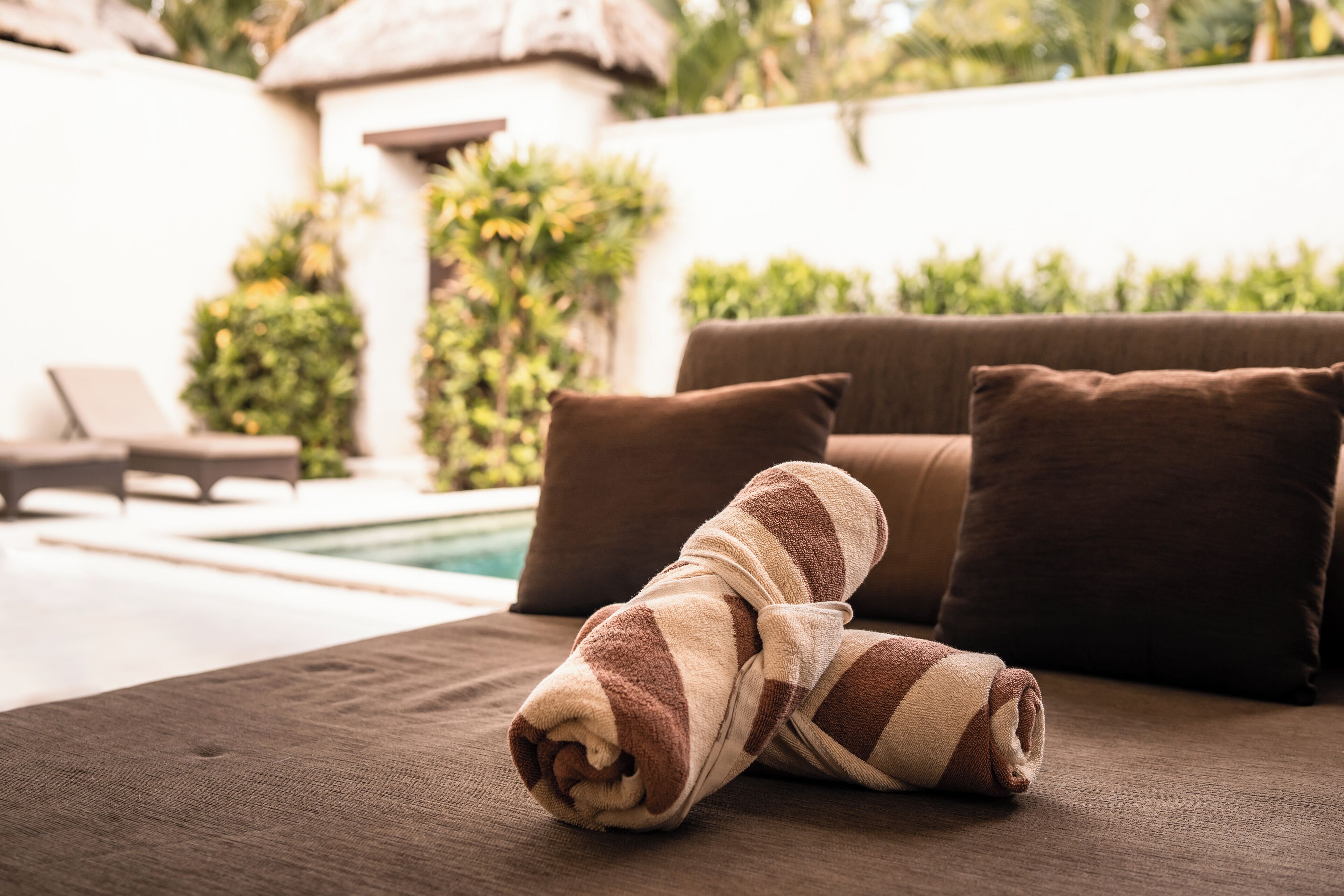 Una cama bien ordenada con toallas a rayas, un sofá marrón con cojines, y una piscina al fondo.