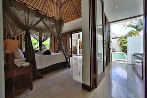 Relax in a serene space with a large bed under a canopy, a bedside table, and a lamp, offering a view of the pool and lush surroundings.