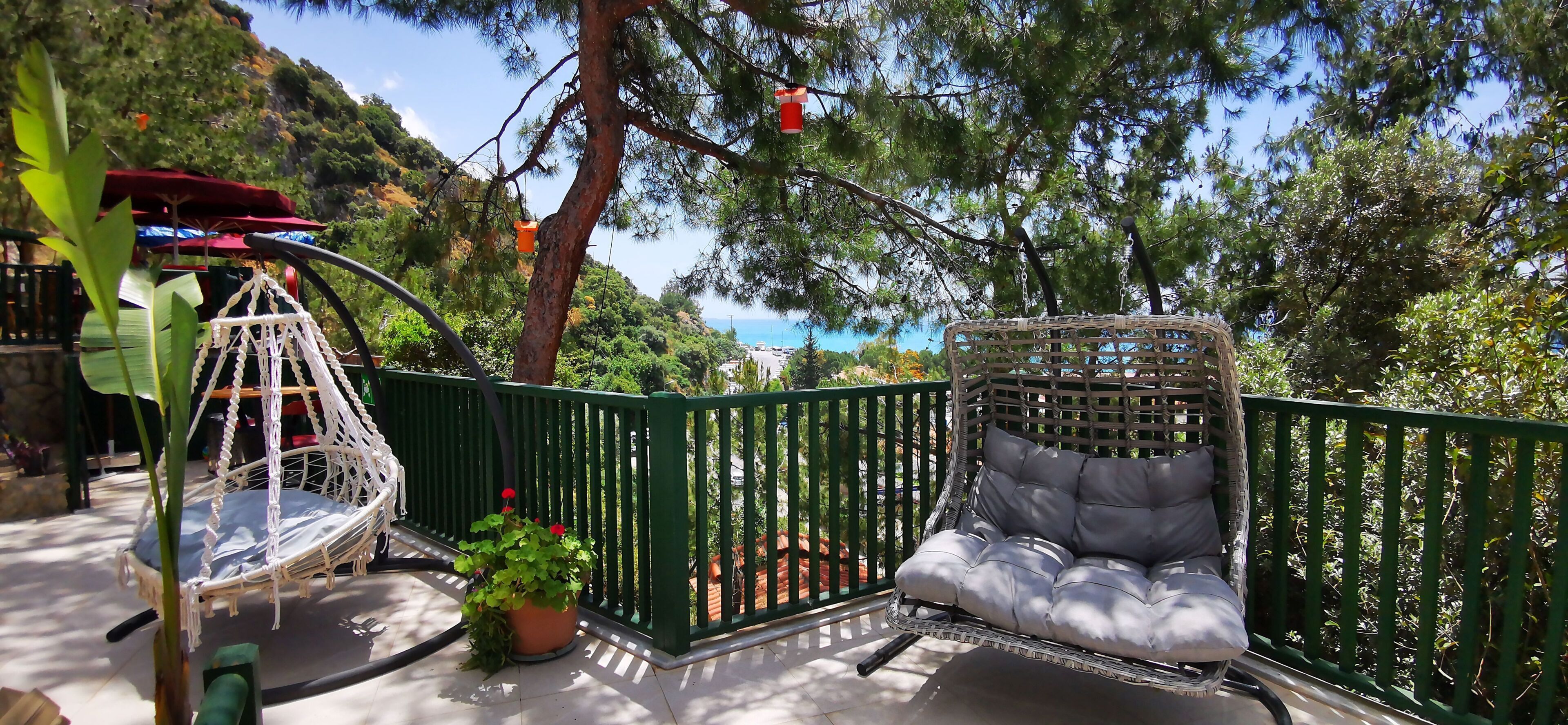 A balcony with a wicker chair and a swing chair, surrounded by a green railing and potted plants.