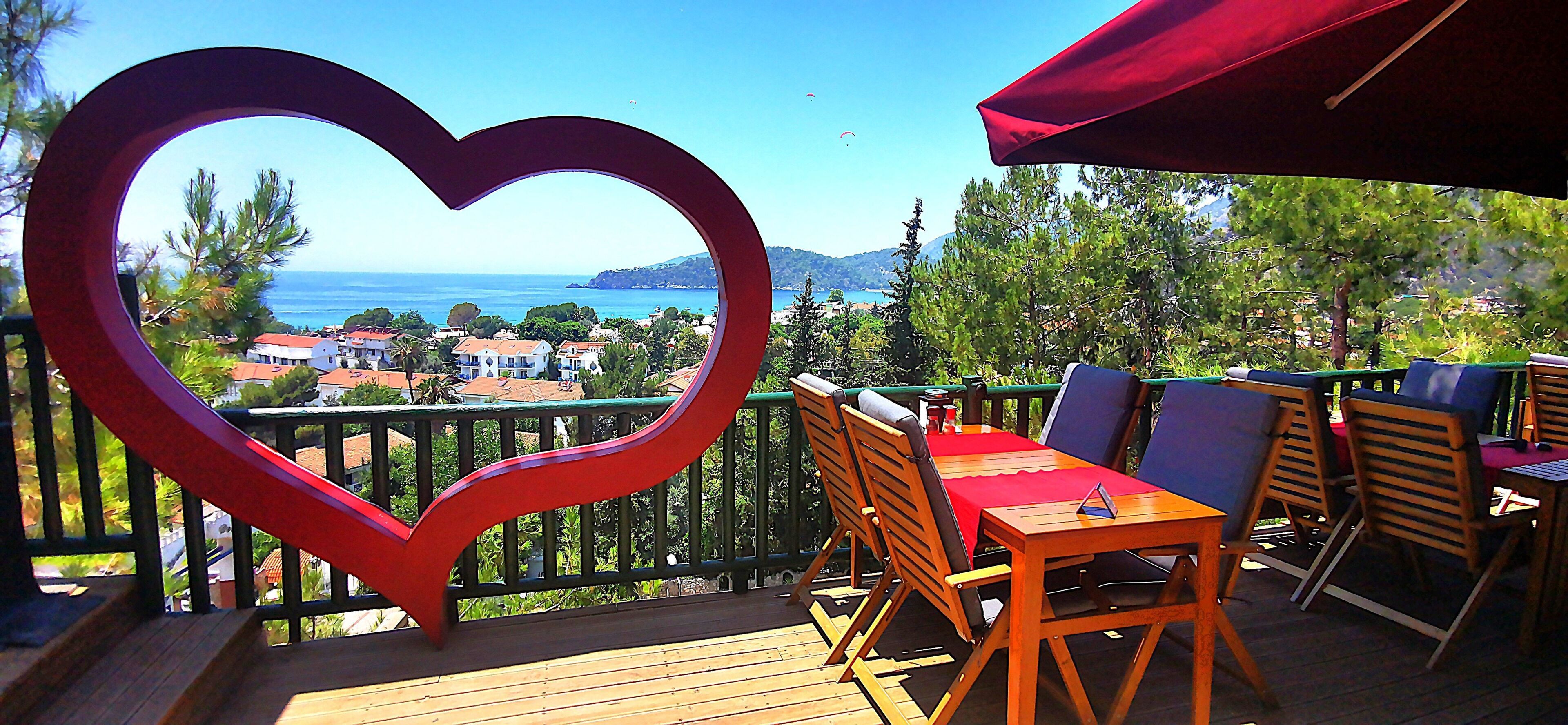 A terrace with a heart-shaped decoration, wooden furniture, and a view of the sea and buildings.