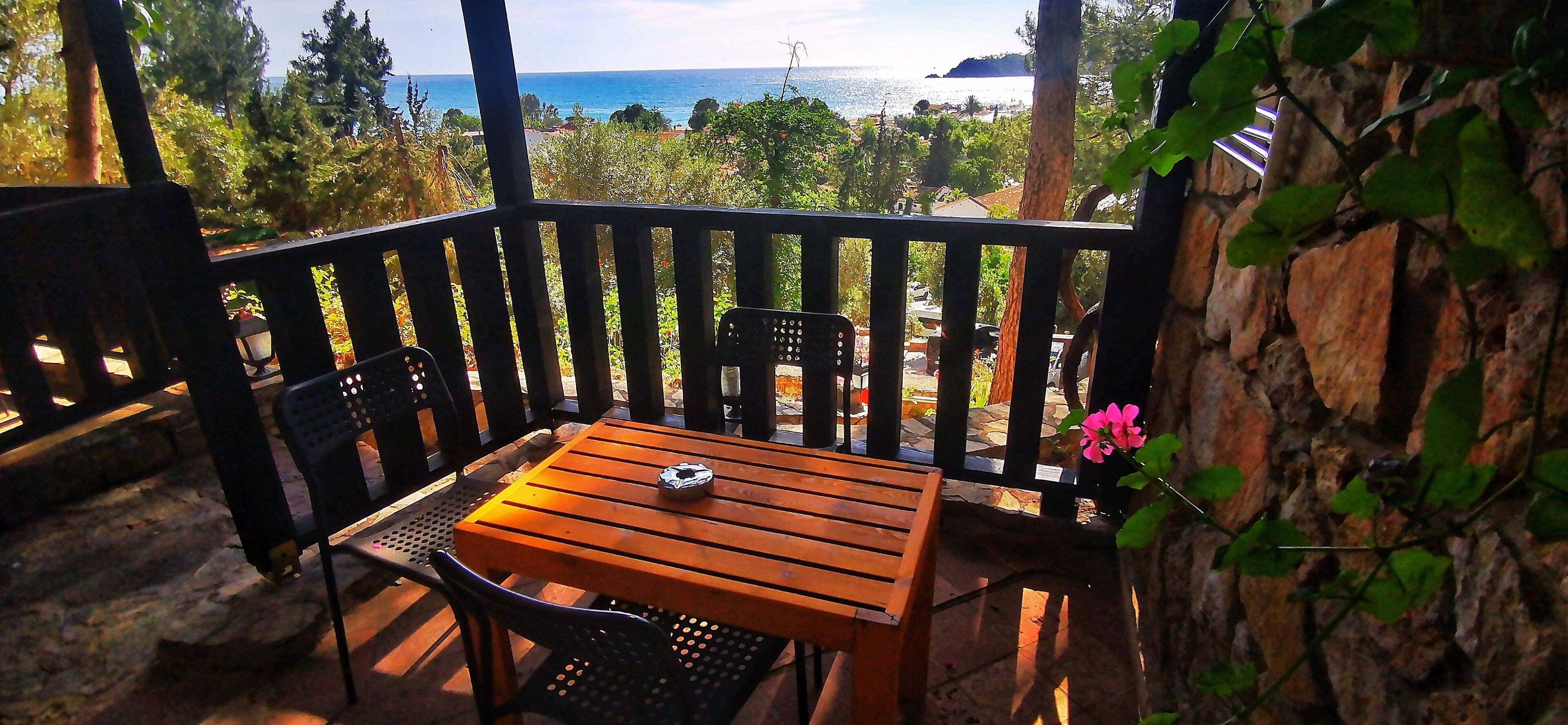 Una terraza de madera con una mesa y sillas, con vistas a un paisaje marino y vegetación.