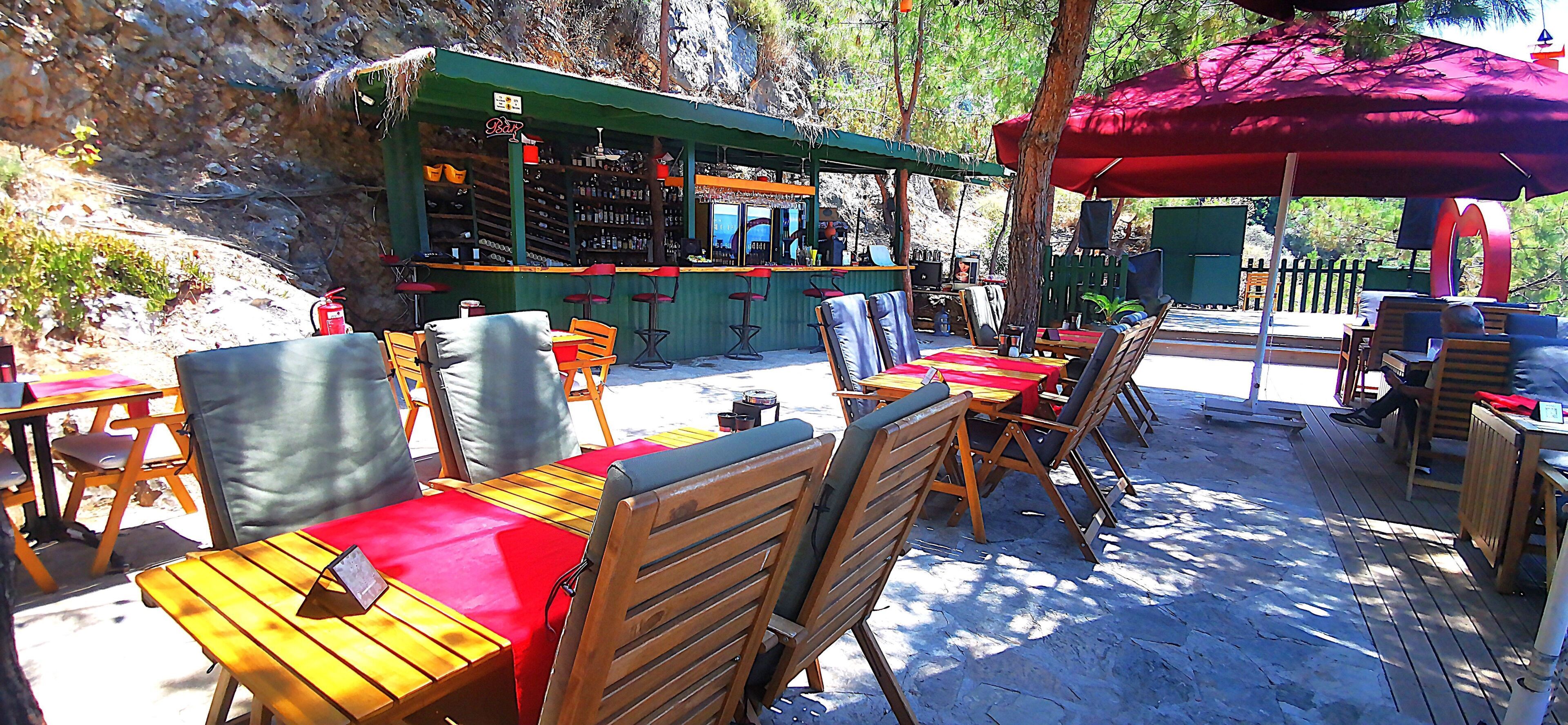 Terras met tafels en stoelen, een bar met flessenplanken, en een rode parasol voor de schaduw.