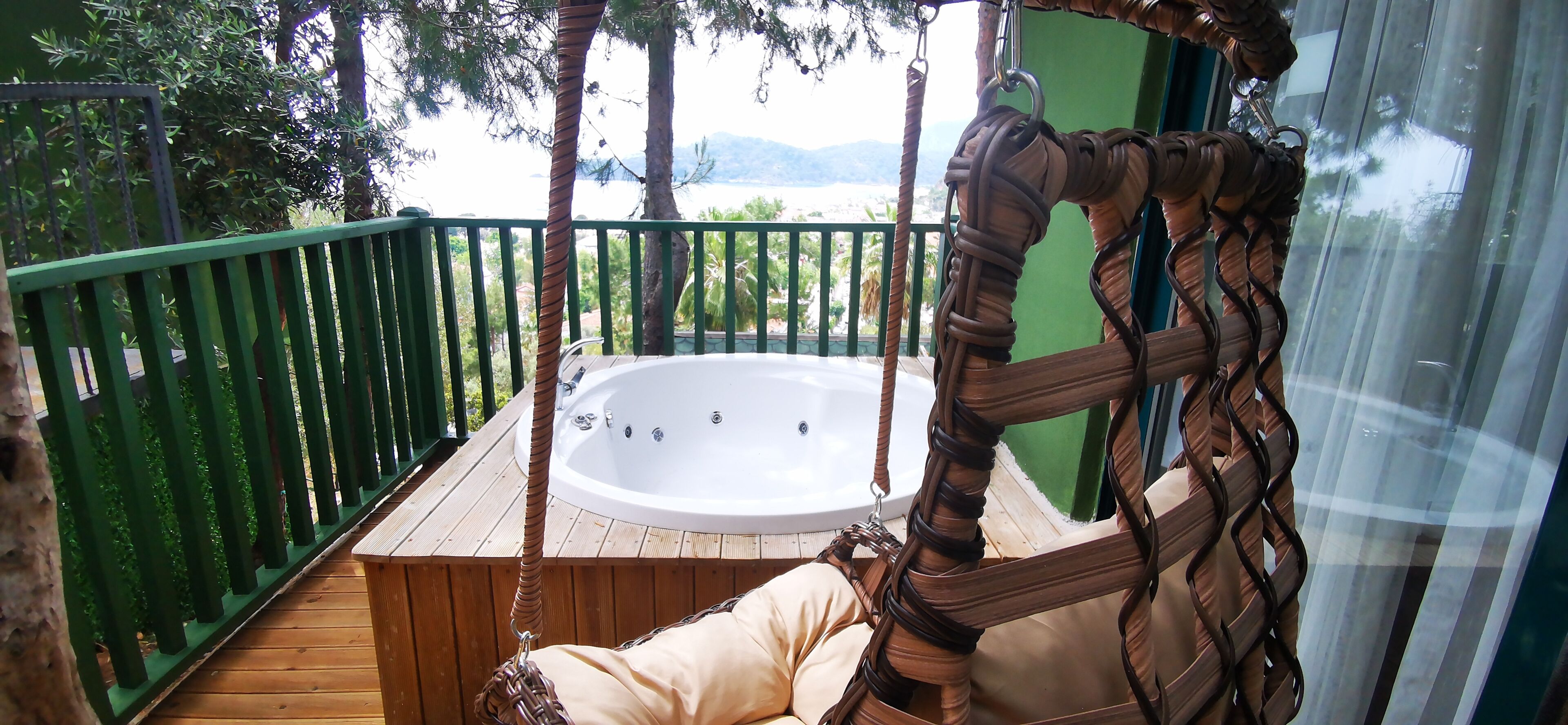 A wooden deck with a hot tub, a wicker swing chair, and a view of the landscape.