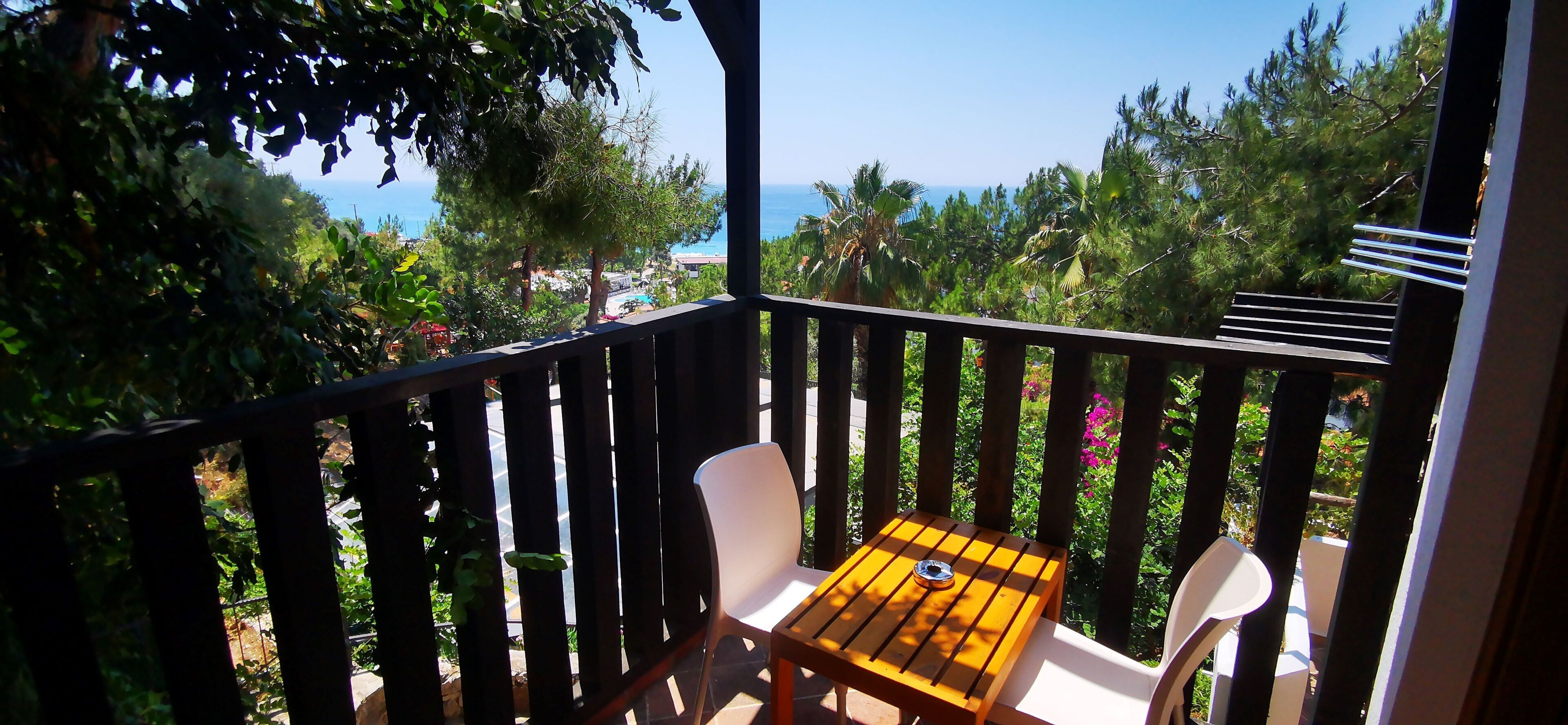 A balcony with a wooden table and two white chairs, overlooking a coastal area with trees and buildings.