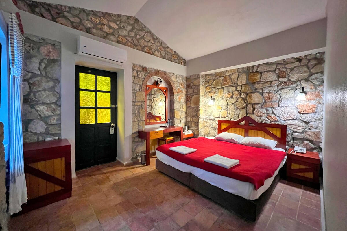 A bedroom with a stone wall, a bed with red bedding, a wooden headboard, a nightstand, a desk, a mirror, and a door with a window.