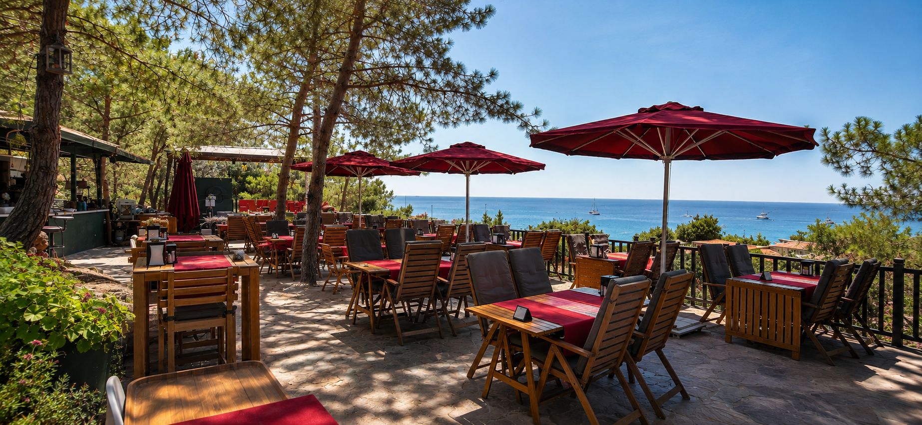 An outdoor seating area with tables and chairs, red umbrellas, and a view of the sea.