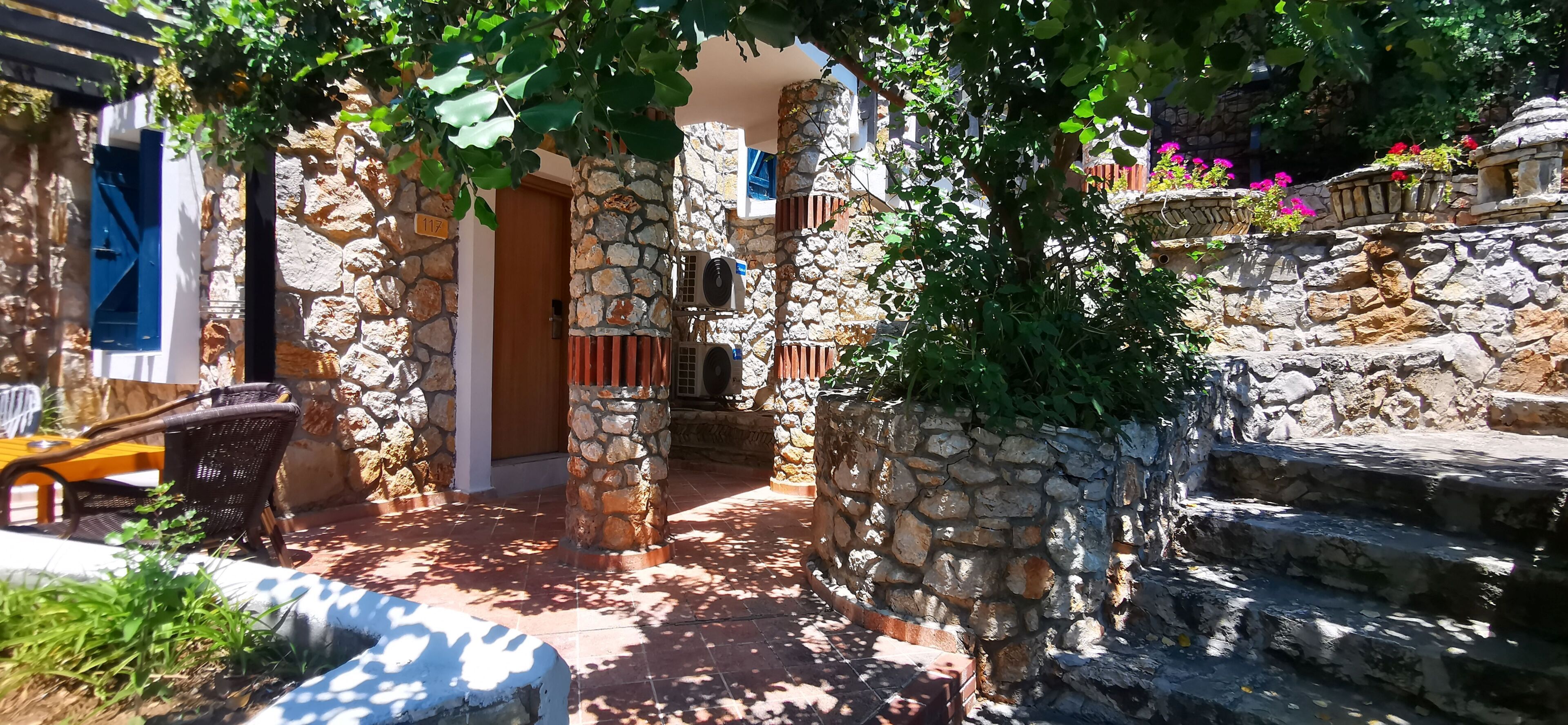 Un patio con paredes de piedra, una puerta de madera, una silla de mimbre y una escalera que sube a un nivel superior.