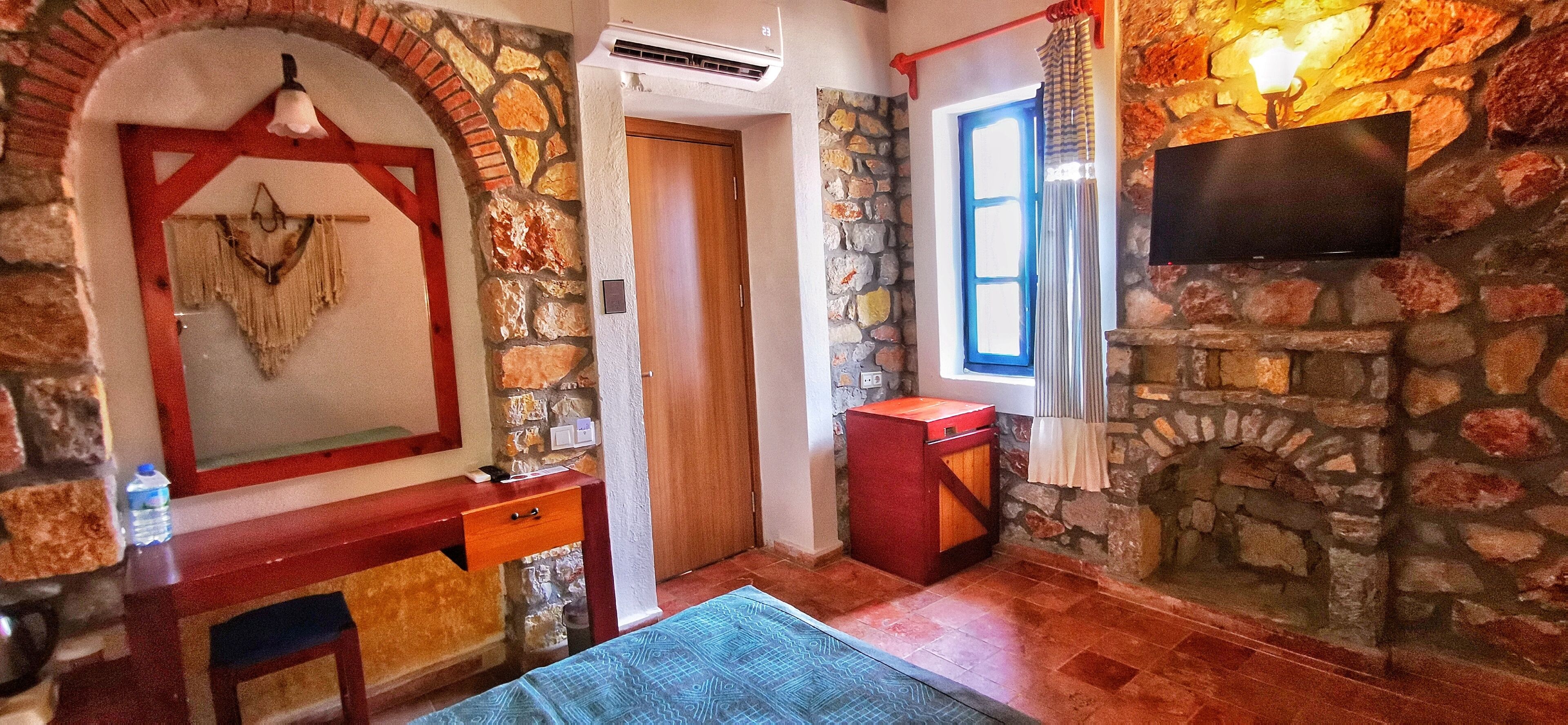 Una habitación con pared de piedra, puerta de madera, televisor montado en la pared y una pequeña mesa con una silla.