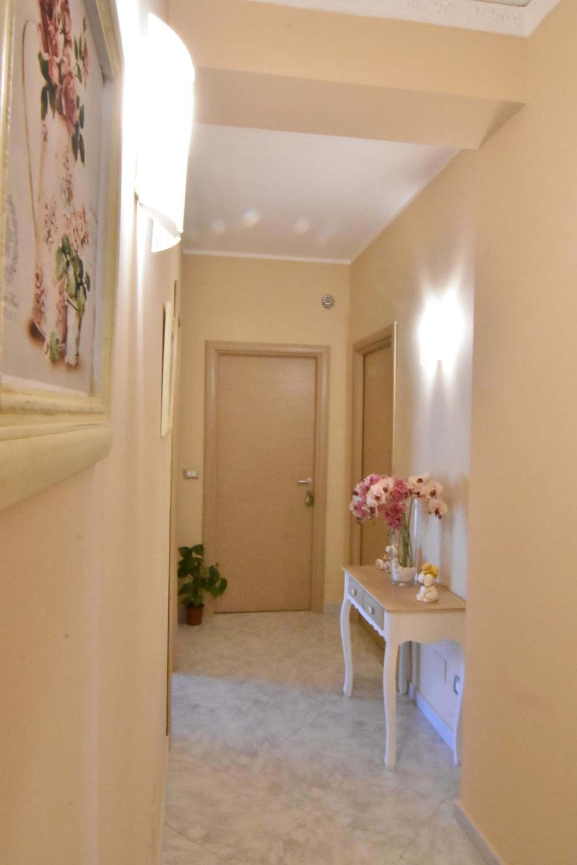 A hallway with a door at the end, a small table with a vase of flowers, and a potted plant.