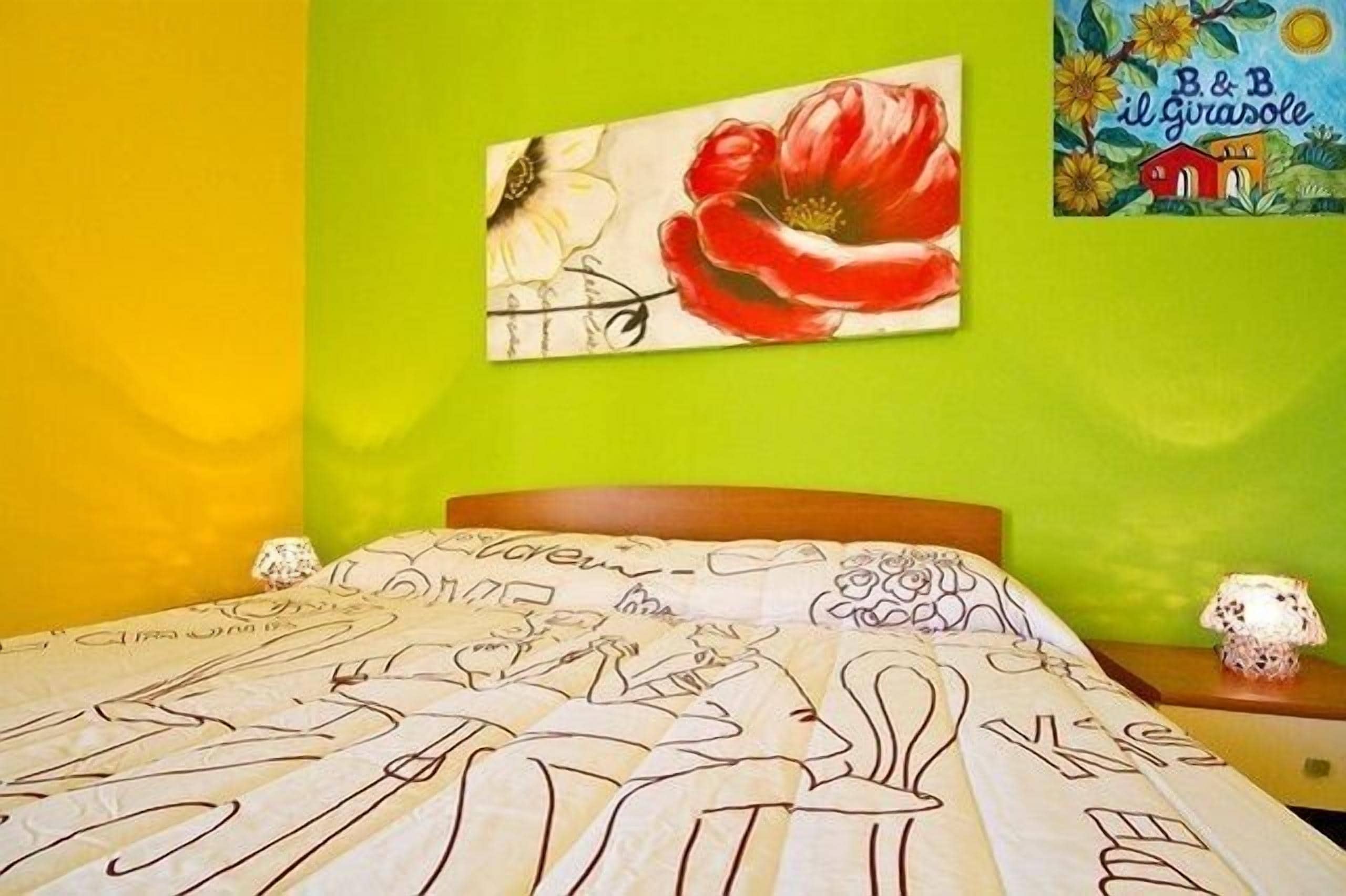 A bedroom with a green wall, a bed with a floral-patterned bedspread, a painting of a red flower, and a book titled 'B&B il girasole'.
