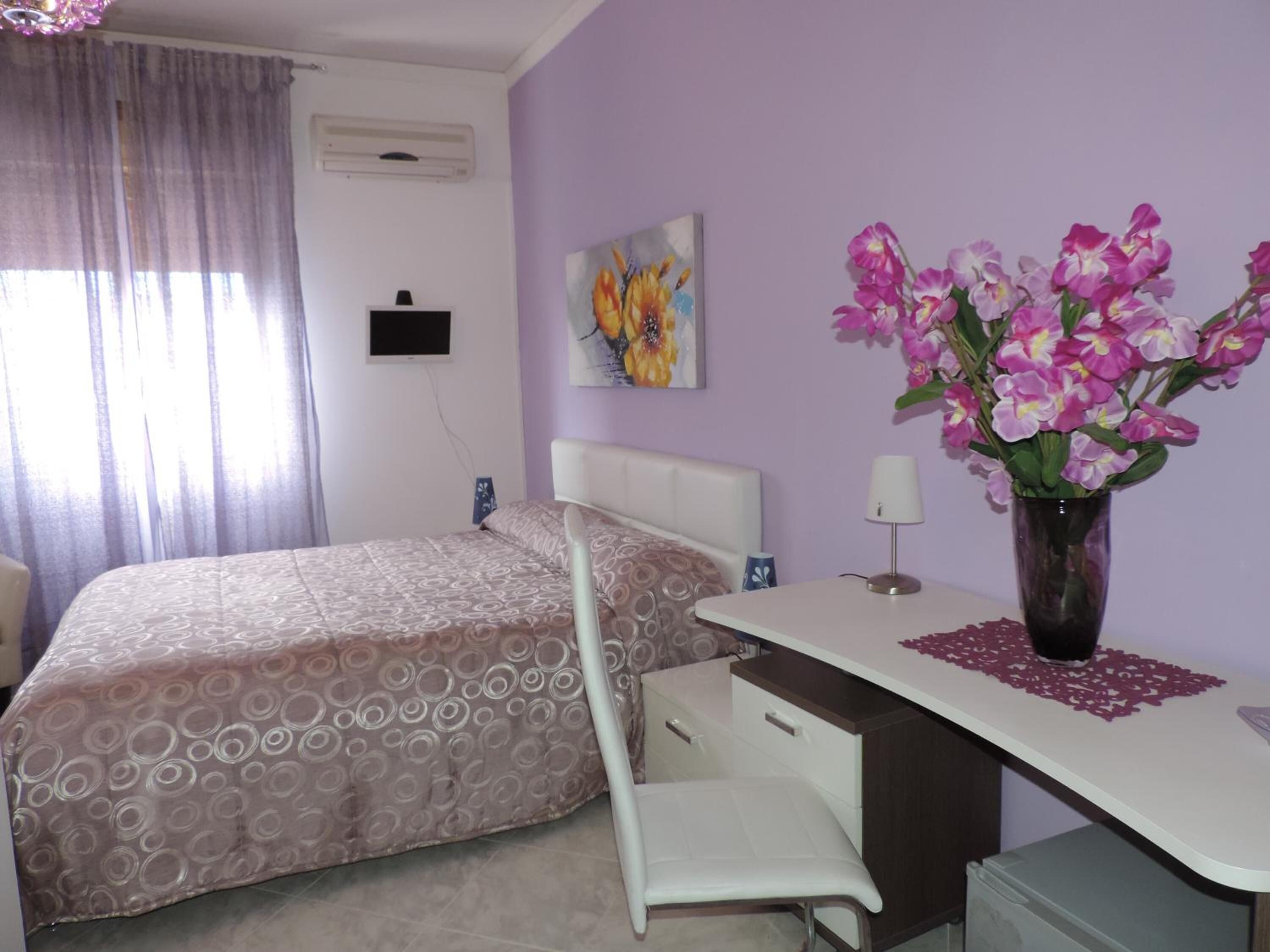 A bedroom with a bed, a desk, a chair, a vase with flowers, and a painting on the wall.