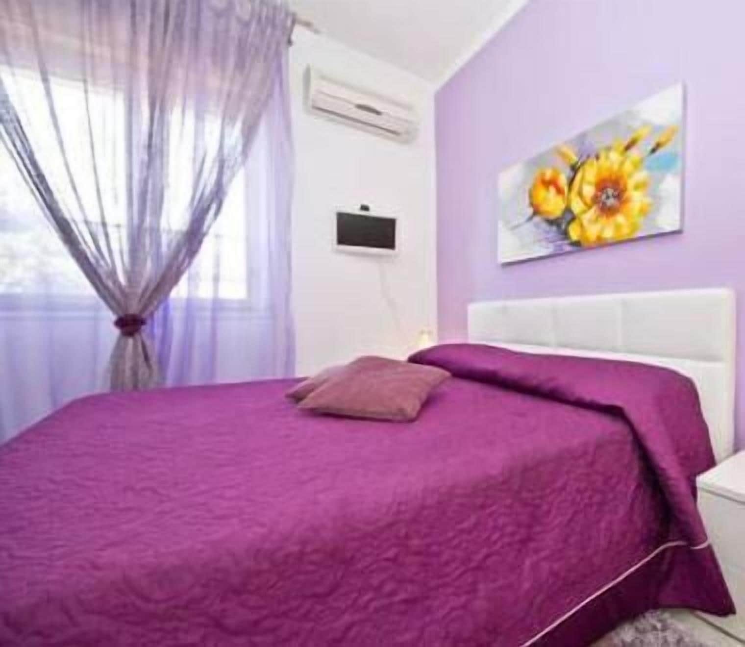 A bedroom with a purple bedspread, a white headboard, and a painting of flowers on the wall.