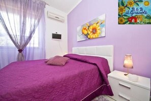 A bedroom with a purple bedspread, a white bedside table with a lamp, and a wall painting. - Il Girasole Bed and Breakfast (Palermo)