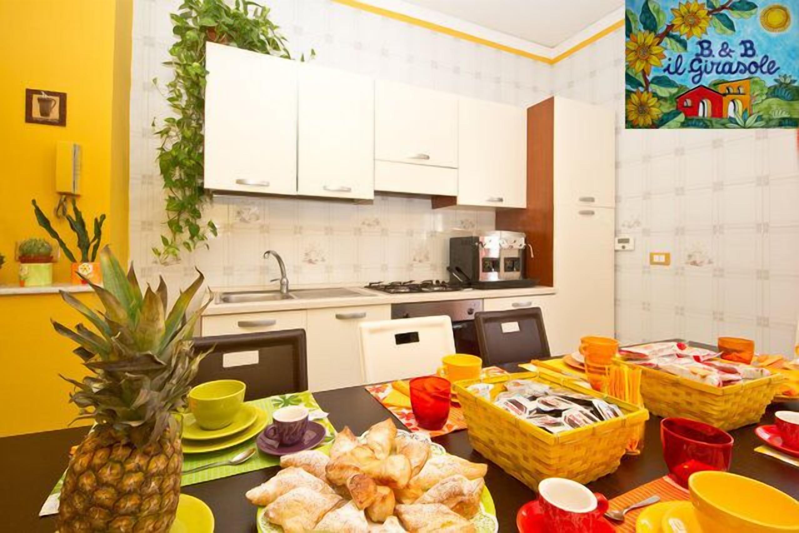Eine Küche mit weißen Schränken, einem Herd und einer Spüle. Ein Tisch ist mit Lebensmitteln und einer Ananas gedeckt.