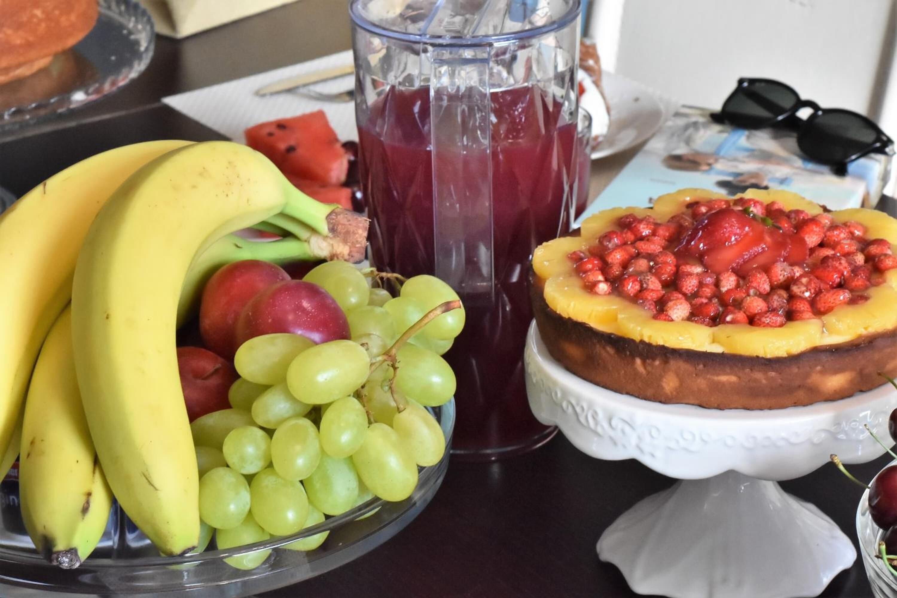 Un assortiment de fruits comprenant des bananes, des raisins et de la pastèque, un verre de jus rouge et un gâteau décoré de fruits, le tout présenté sur un présentoir blanc.