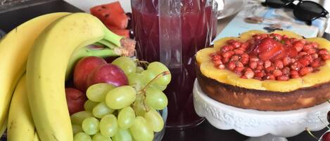 A fruit platter with bananas, grapes, and watermelon, a glass of red juice, and a fruit-topped cake on a white stand.