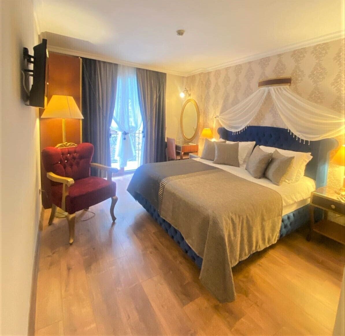 A hotel room with a large bed, a red armchair, a nightstand with a lamp, and a view of the outdoors through a window with curtains.