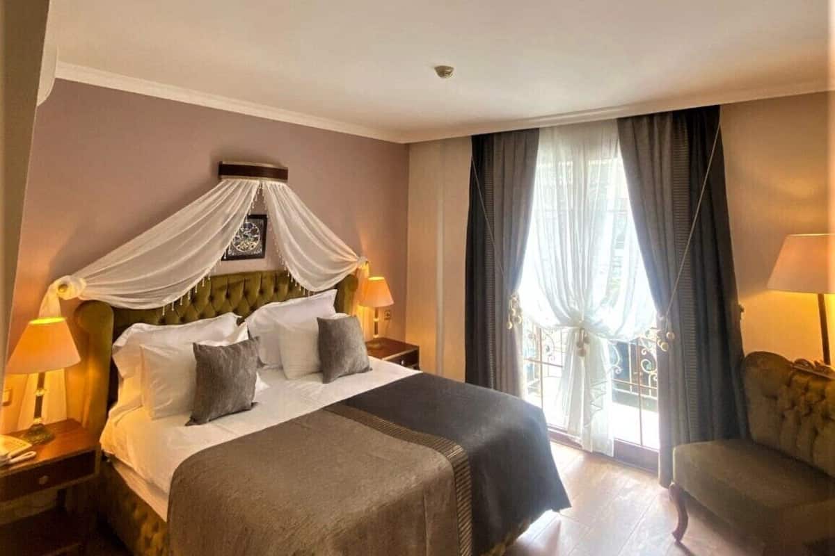 A hotel room with a large bed, a canopy, two bedside lamps, a chair, and a balcony with curtains.
