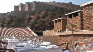 Un comedor con mesas dispuestas para una comida, con vistas a una pared de piedra histórica y una colina con vegetación.